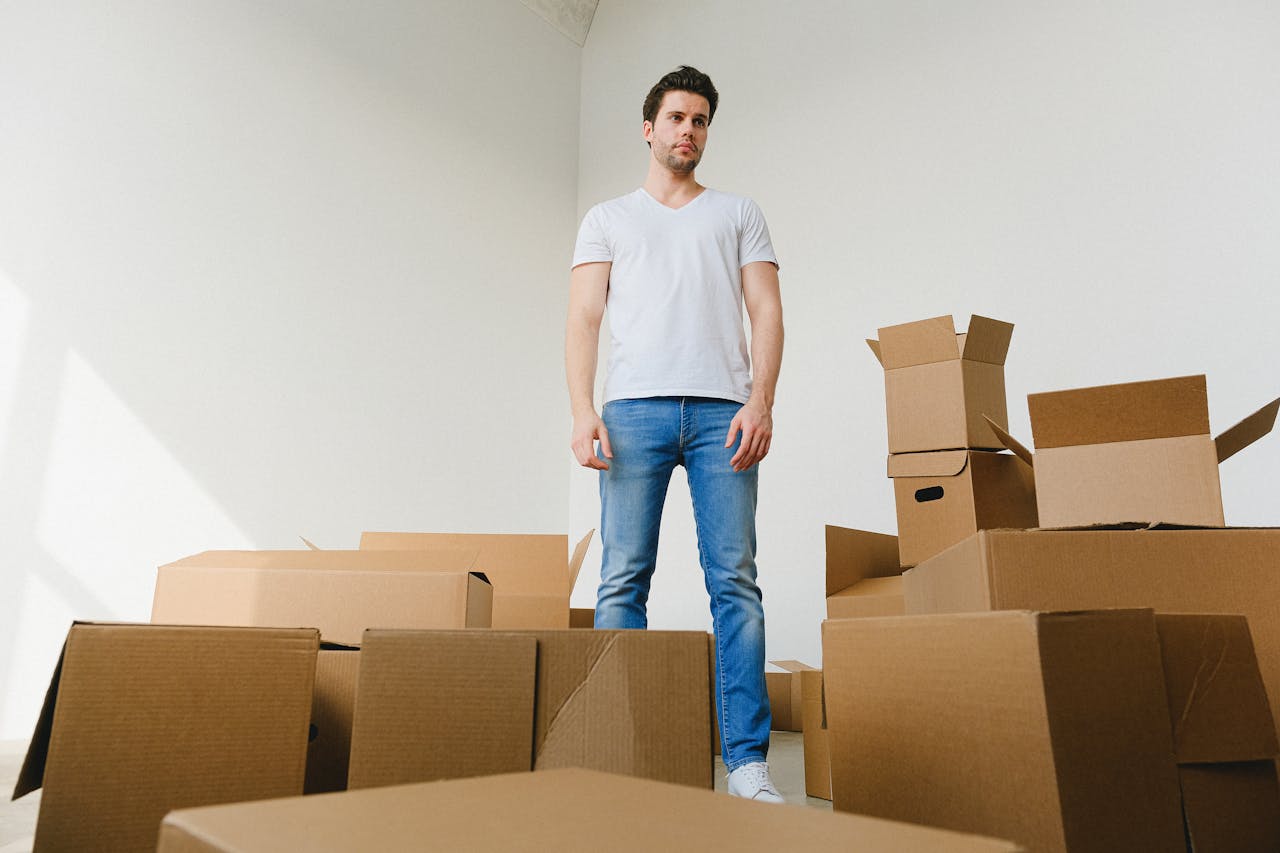 hero-02 Low angle of serious young guy with dark hair in casual outfit standing near pile of cardboard boxes in light room and looking away while preparing for relocation